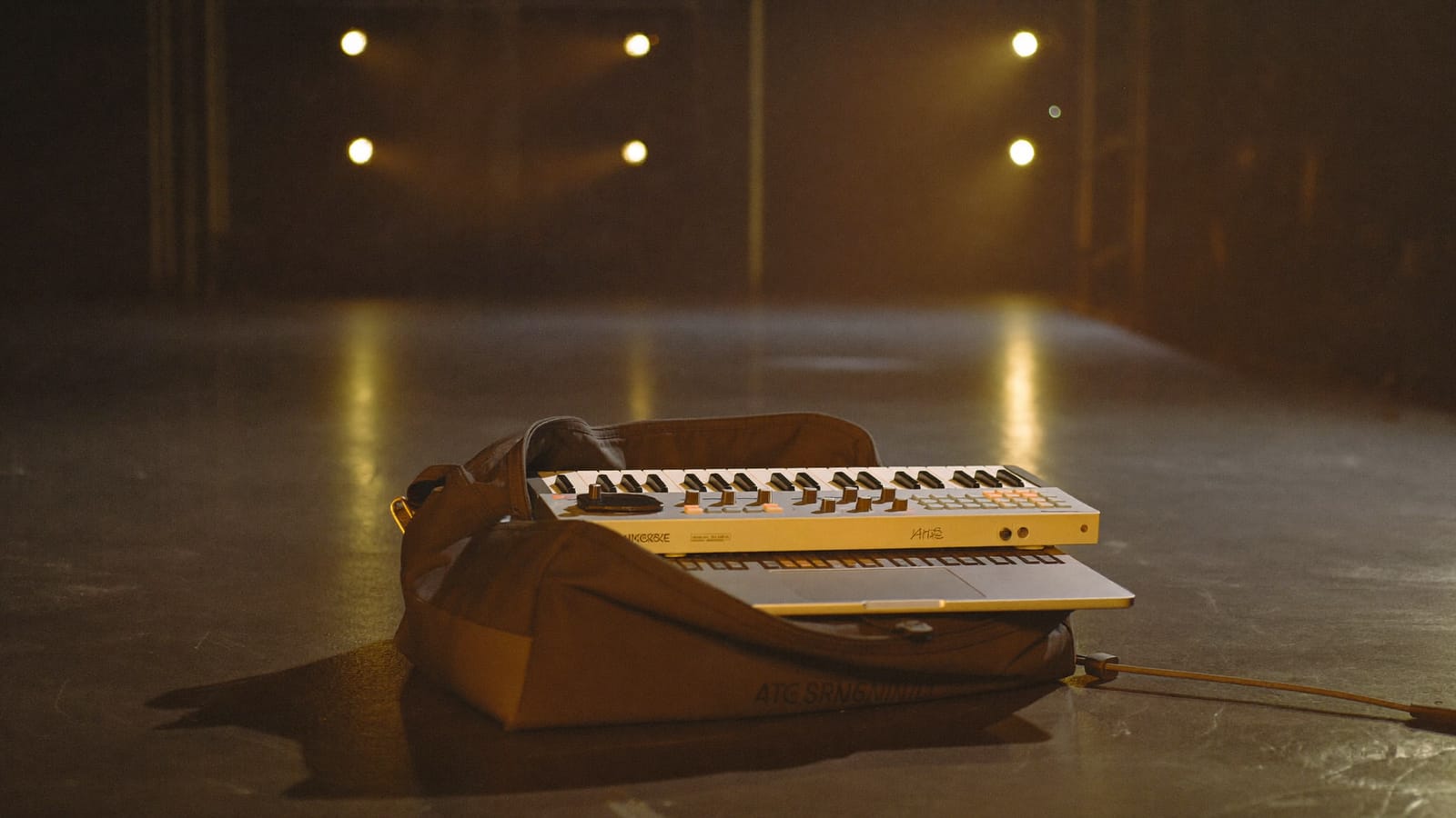 A laptop and compact MIDI controller packed in an open backpack on a dark stage floor under amber light